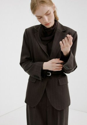 Femme ajustant le poignet de sa veste de costume à fines rayures foncées, portée sur un col roulé noir, debout devant un fond clair uni.