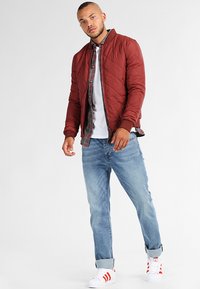 Chaqueta acolchada roja con puños acanalados, sobre una camisa de cuadros y una camiseta blanca, combinada con jeans de mezclilla azules claros y zapatillas blancas.