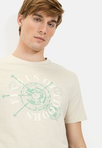 Cremfarbene T-Shirt aus weichem Stoff, mit einem kreisförmigen geometrischen Grafik in Mintgrün und weißem Text in moderner Schriftart.