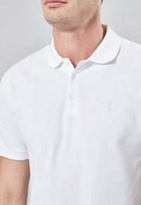 Homme portant un polo blanc à col avec un logo de tête de cerf brodé sur la poitrine, devant un fond gris uni.