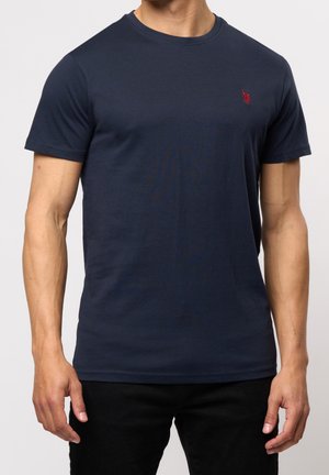 Man wearing plain navy blue short-sleeve t-shirt with small red embroidered logo on left chest, paired with black pants.