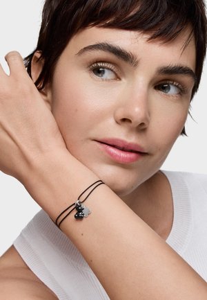 Kobieta o krótkich ciemnych włosach, ubrana w biały bezrękawnik, pokazująca cienką czarną podwójną bransoletkę z małymi srebrnymi i czarnymi zawieszkami.