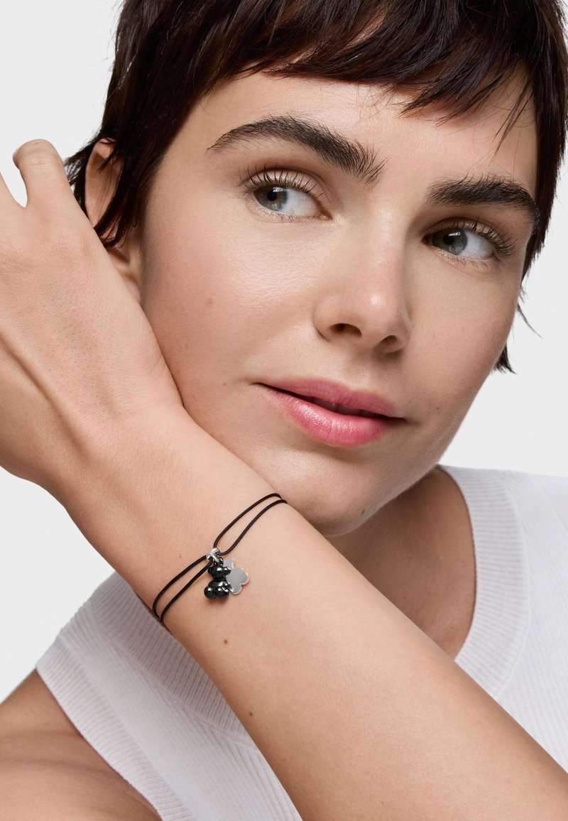 Kobieta o krótkich ciemnych włosach, ubrana w biały bezrękawnik, pokazująca cienką czarną podwójną bransoletkę z małymi srebrnymi i czarnymi zawieszkami.