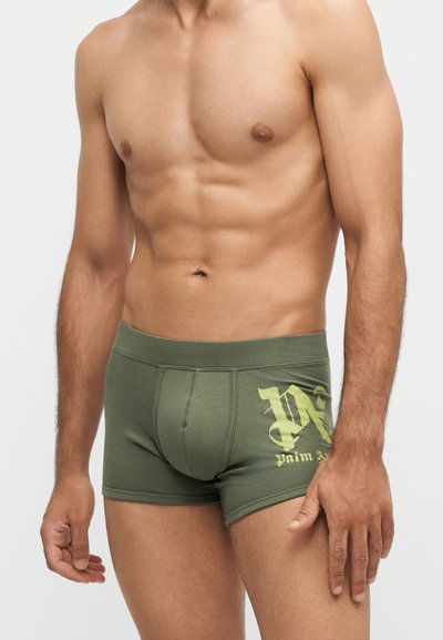 Homme torse nu portant un boxer vert avec le logo jaune "Palm Angels" sur la cuisse gauche, debout devant un fond clair uni.