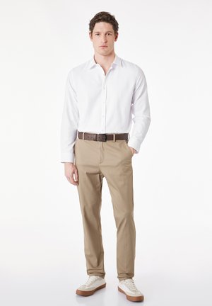 Mann steht mit der Hand in der Hosentasche, trägt ein weißes langärmeliges Hemd mit Knöpfen, beige Hose, braunen Gürtel und weiße Turnschuhe vor weißem Hintergrund.