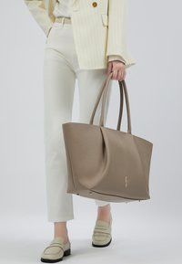 Een persoon in lichte broek, met crème instappers en een gestreepte jas houdt een grote taupe leren tote bag vast bij de hengsels.