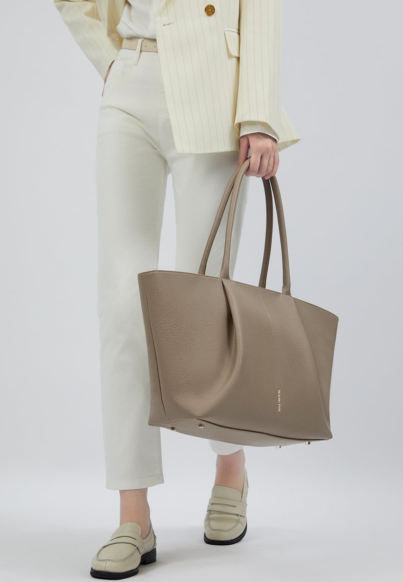 Person iført cremefarvede bukser og loafers holder en stor taupefarvet tekstureret tote-taske med dobbelthåndtag.