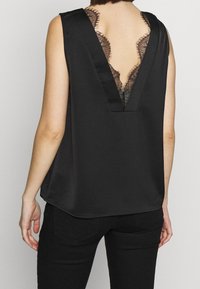 Svart ärmlös topp med djup V-rygg och spetsdetaljer. Slätt satintyg, avslappnad passform och subtila sömnadsdetaljer.