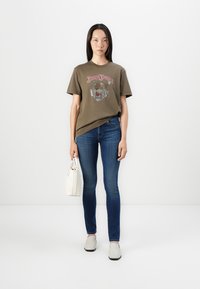 Oliven grønn bomulls t-skjorte med løvegrafikk, kombinert med blå jeans. Hvit håndveske og lysegrå slip-on sko fullfører antrekket.