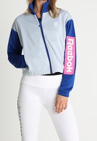 Kvinna iklädd en ljusblå och marinblå zip-up jacka med rosa Reebok-logga på ärmen, i kombination med vita leggings som har svart text på låret.