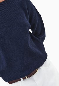 Granatowy sweter robiony na drutach z dekoltem off-shoulder, teksturowana tkanina i ściągacz u dołu, w połączeniu z brązowym paskiem i białymi spodniami.