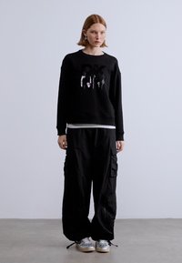 Μαύρο φούτερ με λογότυπο, συνδυασμένο με φαρδιά μαύρη φόρμα cargo με πολλαπλές τσέπες. Το μοντέλο φοράει ασημί αθλητικά παπούτσια.