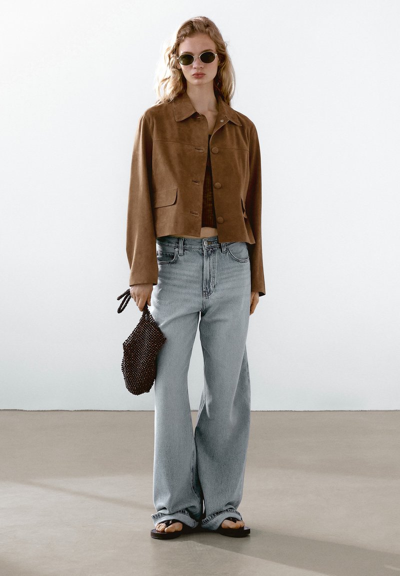 Giacca corta in suede marrone con bottoni frontali, jeans a gamba larga blu chiaro, borsa in perline nera e occhiali da sole rotondi scuri. Tessuto morbido.