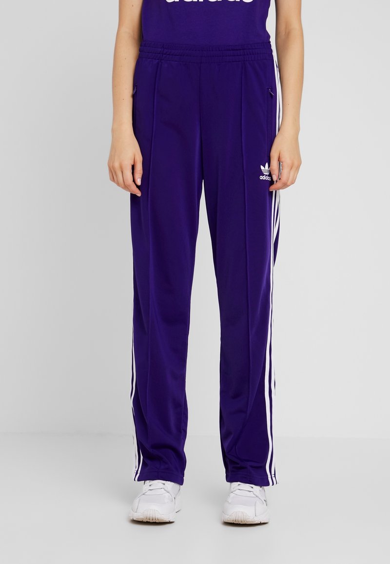 Lila träningsbyxor gjorda av lättviktig tyg, med vita sidostraper och en subtil logga på höften, samt ett elastiskt midjeband.