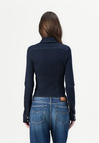 Mørk denimjakke med en tettsittende design, lange ermer og krage. Har knappede mansjetter og et skinnmerke på midjen.