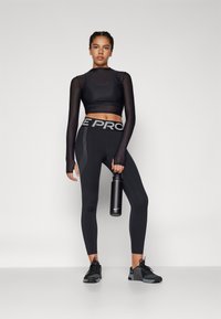 Top crop negru cu mâneci lungi transparente, tigăi negre cu talie înaltă și logo reflectorizant, cu accente texturate. Ținând o sticlă de apă neagră.