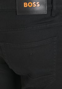 Svarta jeans i denim med en slät textur, med en läderetikett med "BOSS" i orange och en detalj på bakfickan.