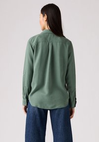 Groen overhemd met lange mouwen van gladde stof, voorzien van een puntig kraag, knoopmanchetten en een rugplooi. Gecombineerd met spijkerbroek.