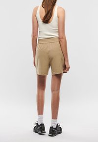 Woman standing facing away wearing beige shorts, white sleeveless top, white socks, and black sports shoes on a plain white background.