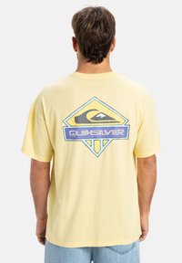 Žluté bavlněné tričko s velkým logem Quiksilver v modré a černé barvě umístěným v designu v tvaru diamantu na zádech. Krátké rukávy, volný střih.