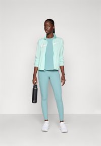 Akvamarinfärgad atletisk klädsel bestående av en lätt jacka, en åtsittande topp och leggings. Tillbehör inkluderar en svart vattenflaska och vita sneakers.