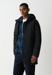 Juodas gobtuvas striukė su lygiu išoriniu paviršiumi, dėvima virš mėlynos užtraukiamos flisinės striukės. Yra šoninės kišenės ir tiesus siluetas.