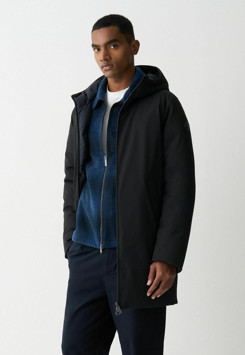 Juodas gobtuvas striukė su lygiu išoriniu paviršiumi, dėvima virš mėlynos užtraukiamos flisinės striukės. Yra šoninės kišenės ir tiesus siluetas.