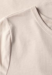 Un t-shirt beige à manches courtes en coton doux et lisse. Il possède un col rond et des coutures visibles le long des bords.