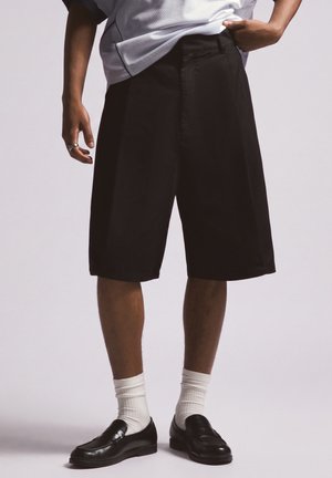 Person trägt schwarze lange Shorts, weiße Socken, schwarze Loafer und ein weißes Hemd, steht mit einer Hand in der Tasche vor einem einfarbigen Hintergrund.