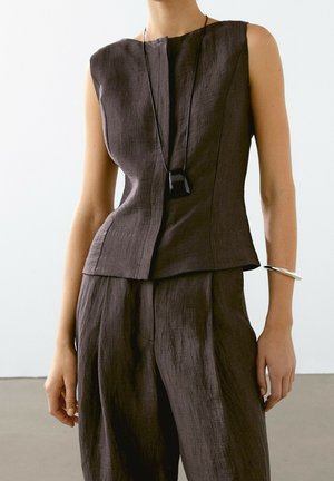 Sleeveless brown linen top and matching wide-leg pants worn by a model, accessorized with a black pendant necklace and silver bangle bracelet.