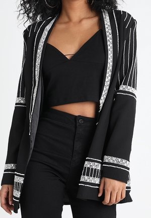 Vrouw draagt een zwarte cropped top met een zwarte broek met hoge taille en een zwart colbert versierd met wit kralenpatroon.