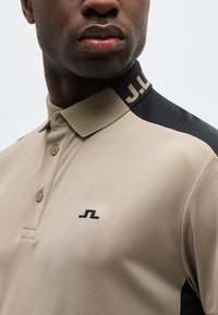 Άνδρας που φοράει μπεζ πόλο πουκάμισο με μαύρες πανελ στους ώμους και μικρό μαύρο λογότυπο στο στήθος, καθώς και επωνυμία στο γιακά.