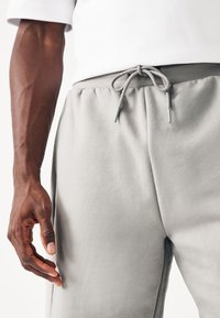 Homme portant un pantalon de survêtement gris clair avec un cordon noué et une chemise blanche, montrant son bras gauche et sa main détendus le long du corps.