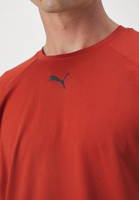 Vyras, vilkintis raudoną Puma marškinėlį su juodu Puma logotipu ant krūtinės, nukirptas apie kaklą ir pečius, prieš vienodą fono vaizdą.