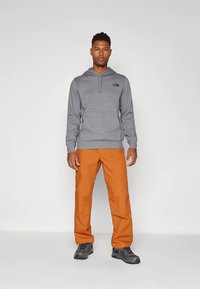Sweatshirt cinzento com capuz e logótipo no peito, combinado com calças laranja e sapatos de caminhada pretos, em pé sobre um fundo simples.