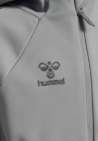 Graue Sportjacke mit glatter Stofftextur, ausgestattet mit einem zentralen Reißverschluss und einem schwarzen, geprägten Logo im Brustbereich.
