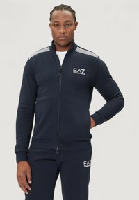 Marinblå zip-up träningsjacka i mjukt material med hög krage, med vit logotyp och randiga axeldetaljer. Slät textur, figurnära design.