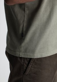 Zijaanzicht van een persoon in een olijfgroen t-shirt en donkerbruine broek, met nadruk op de onderkant van het t-shirt en het zakgebied van de broek.