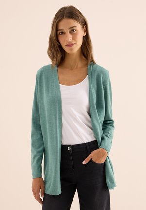 Jeune femme portant un cardigan léger de couleur teal clair, un top blanc à col bateau et un pantalon noir, se tenant devant un fond beige.
