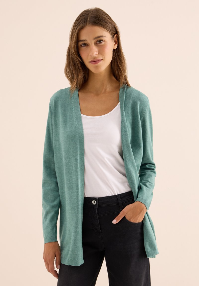 Mladá žena v světle tyrkysovém otevřeném svetru, bílém topu s kulatým výstřihem a černých kalhotách, stojící na béžovém pozadí.