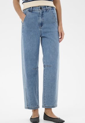 Personne portant un jean taille haute, bleu clair, coupe droite, avec une main dans la poche et des ballerines noires à pois blancs sur fond blanc.