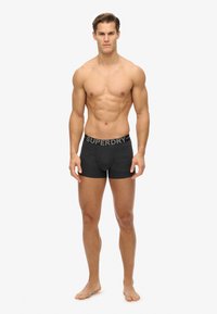 Zwarte katoenen boxershorts met een tailleband waarop het logo "SUPERDRY" staat. Model staat blootsvoets, toont een getraind figuur en heeft een neutrale uitdrukking.