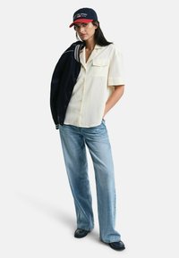 Creme kortærmet skjorte, lyseblå jeans, marineblå jakke over skulderen, rød og marineblå kasket med tekst. Flade sko, afslappet pasform i bukser, afslappet stil.