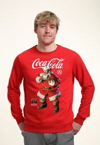 Červená mikina s logem Coca-Cola a grafikou Santa Clause. Měkká tkanina s dlouhými rukávy a žebrovanými manžetami.