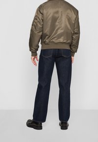 Oliemgroen satijnen bomberjack met geribbelde zoom, gecombineerd met donkerblauwe rechte jeans en zwarte leren laarzen, gezien van achteren.