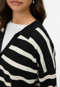 Cardigan à rayures noir et blanc avec col, présentant une texture en tricot douce. Les rayures varient en largeur et contrastent avec un fond uni.