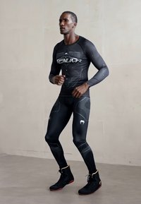 Mężczyzna w czarnym odzieży kompresyjnej sportowej i wysokich butach sportowych biega w pomieszczeniu na tle jasnej ściany.