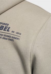 Felpa beige con texture a coste. Presenta testo blu scuro con la scritta "DEFINED LABEL RR - 2013" e "FINNISH DESIGNED GARMENTS."