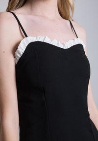 Rochie neagră cu un design ajustat, bretele subțiri și un decolteu alb cu volănașe. Textură netedă cu detalii ale cusăturilor curbe.