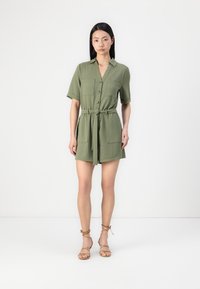 Robe olive verte à manches courtes avec fermeture à boutons, deux poches poitrine et ceinture à la taille. Texture douce avec une coupe confortable. Chaussures : sandales à lanières beiges.